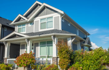 A modern, two-story house with gray siding, white trim, and a front porch. It is surrounded by plants and bushes, under a clear blue sky.
