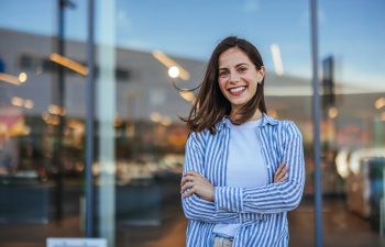 A woman with long brown hair smiles with arms crossed, standing in front of a glass building entrance. She is wearing a white shirt and a blue-and-white striped shirt.